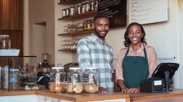 Portrait of two smiling young African entrepreneurs standing welcomingly together behind the counter of their trendy cafe
